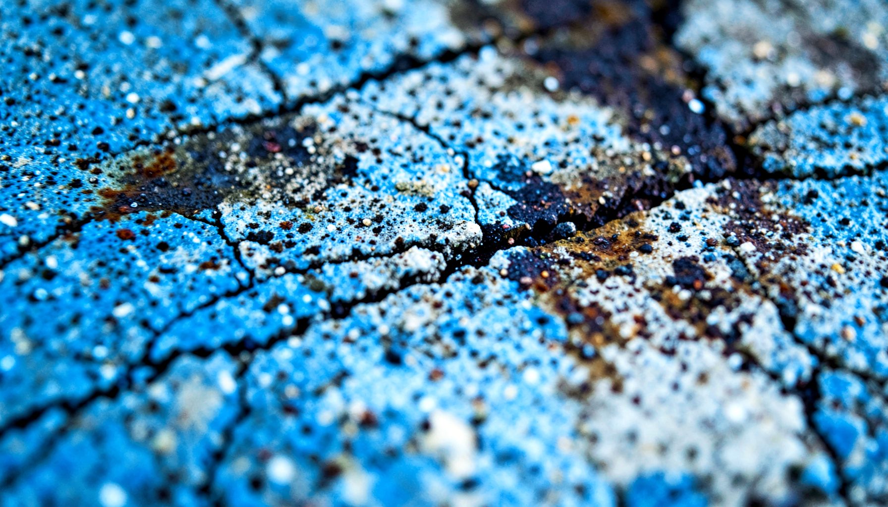 Extreme close-up of cracked, weathered industrial concrete with dark staining and rust-colored residue, gritty texture