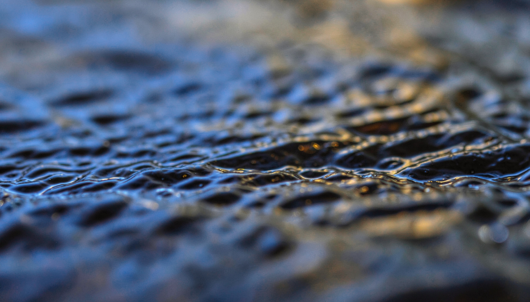 Close-up of oily sheen on dark water surface with ripples, muted gray and amber tones