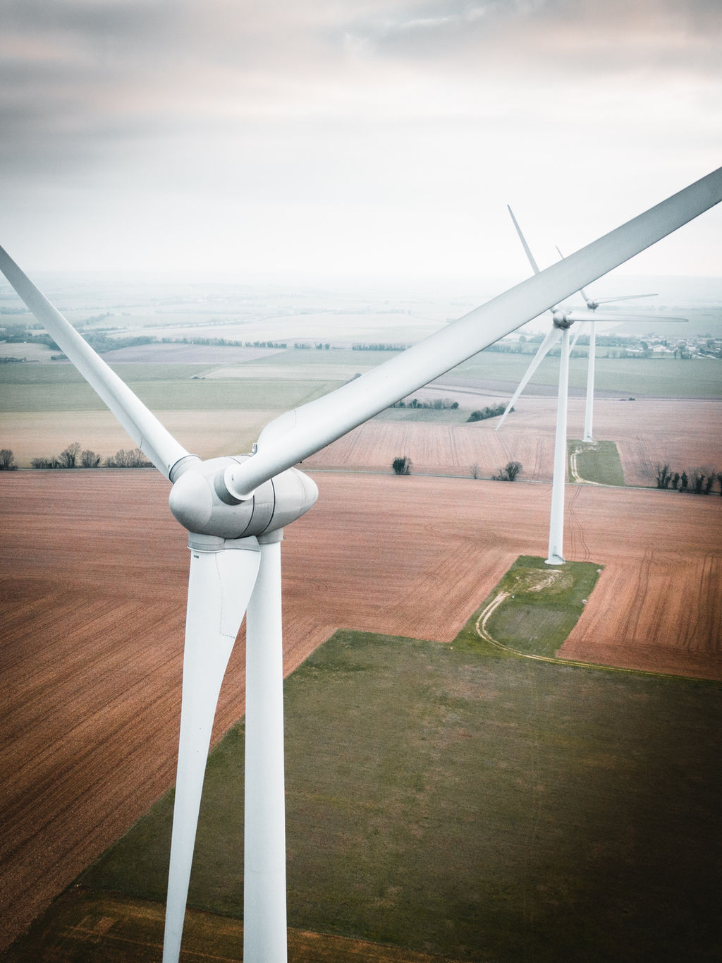 Aerial view of wind turbines over farmland with patchwork fields in brown and green tones, overcast sky, calm industrial